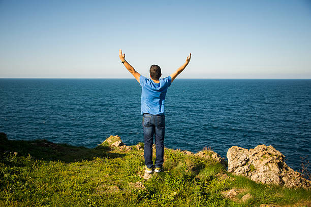 A young man stands with arms wide open in front of the sea.