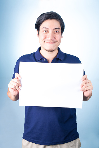 Asian handsome man standing and showing empty horizontal blank paper in hands, on blue background