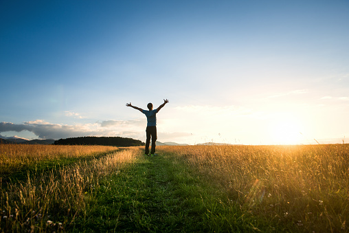 Male silhouette raising his hands and standing on a sunlit meadow at sunset