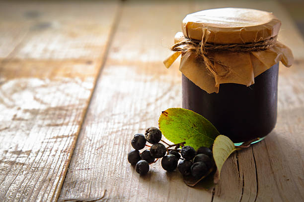 Glass jar of chokeberry jam and fresh berries with few leaves on rustic wooden table.