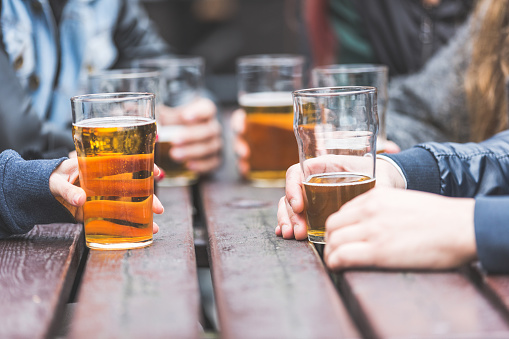 Hands holding glasses with beer on a table at pub in London. A group of friends is enjoying beer time in the city, close up on the glasses.