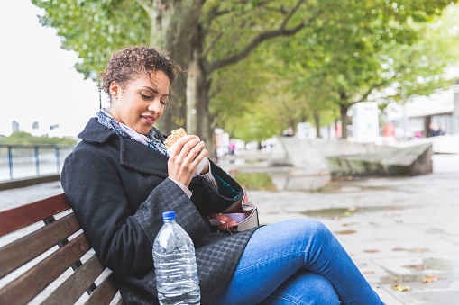 Young woman having lunch in London. She is sitting on a bench, eating a sandwich, typical lunch for many workers in the city. Urban lifestyle concept.