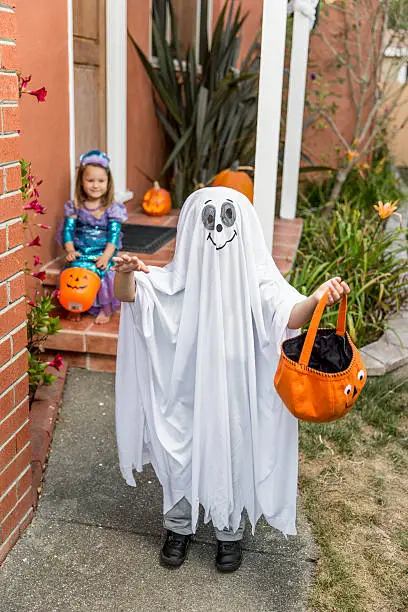 A smiling ghost and a little princess on the front porch with a treat bag on Halloween.