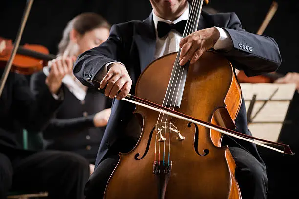 Cello professional player with symphony orchestra performing in concert on background.