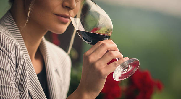 Winetasting. Closeup of unrecognizable adult woman holding a glass of red wine and smelling it before tasting. She's standing outdoors on summer afternoon. Blurry gras and red flowers in background. Toned image. tasting stock pictures, royalty-free photos & images