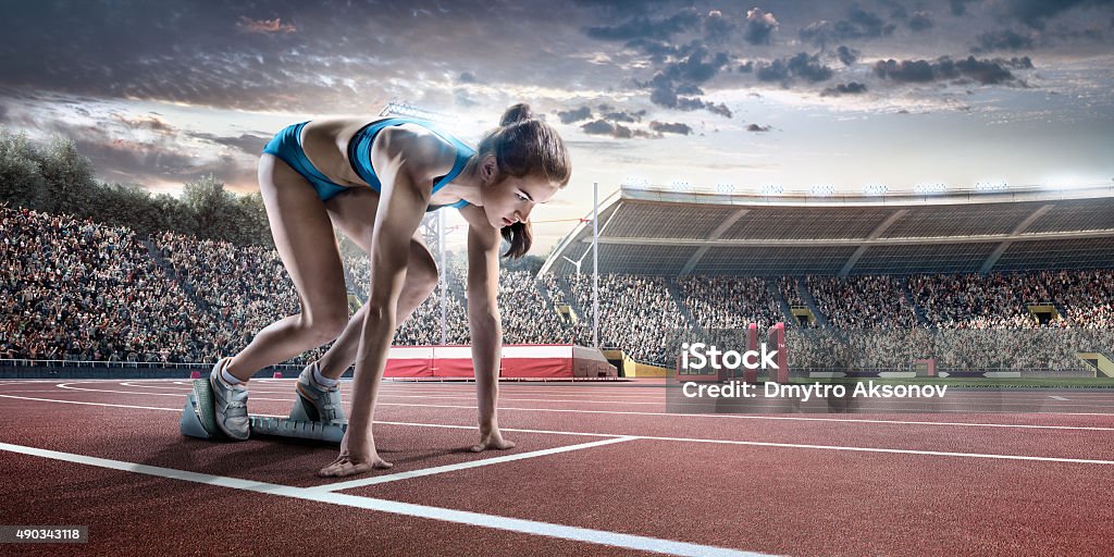 Female athlete prepares to run Professional female athlete sprinting from blocks on numbered start line on outdoor athletics track on . stadium full of spectators under a dramatic evening sky. Sprinter is wearing generic athletics kit. American Football - Sport Stock Photo