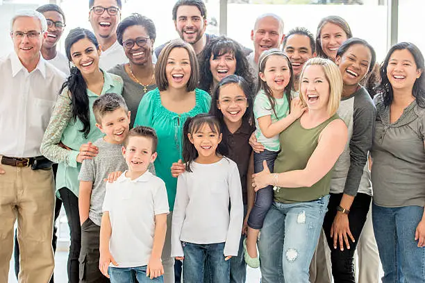 A multi-ethnic and multi-generational group of people standing together, they are smiling and laughing while looking at the camera.
