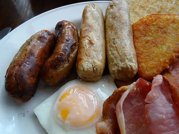 Photo showing some pork and vegetarian sausages on a plate with bacon, a fried egg and hash browns - a full English fried breakfast.