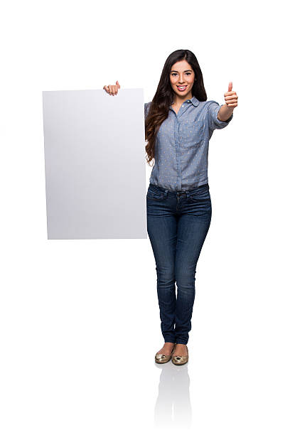 Full length / looking down / front view / one person of 20-29 years old adult beautiful redhead caucasian female / young women standing wearing double denim / jeans / pants / denim jacket / boot / rolled-up sleeves / jacket / warm clothing who is smiling / happy / cheerful / laughing who is pointing / showing and holding sign / banner - sign and using placard with copy space