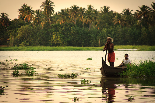 Kottayam (Kerala), India - November 24, 2009: A fisherman standing and poling in his traditional canoe in a lagoon in the Southern Kerala backwaters at dusk. A child sits behind him.