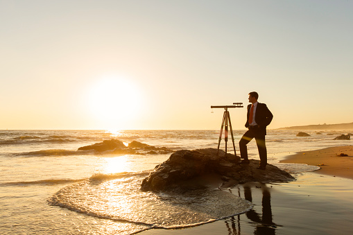 A young businessman in suit stands on a California beach during sunset hoping for a great business future. His telescope sits on a rock while the surf rolls in and the sun sets on the horizon.