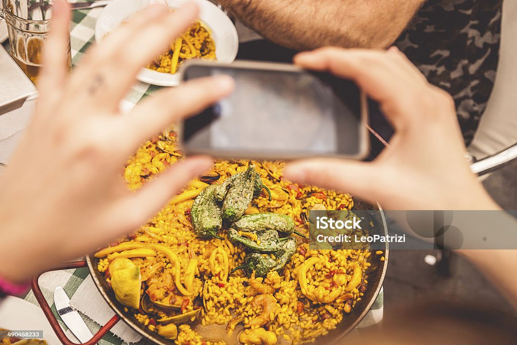 Taking a photo of Spanish paella on smartphone A photo of two hands holding a camera and taking a picture of Spanish paella.  The hands and camera are out of focus while the paella and surrounding items are in focus.  The left hand is coming from the left side of the image, and the right hand is coming from the right side of the image with the camera in the top middle.  In a metal dish, the paella includes green peppers, shrimp, rice, octopus and clams.  A short drinking glass on the table is in the upper left corner of the image.  An arm with a small bowl of paella is at the top of the photo. Adult Stock Photo