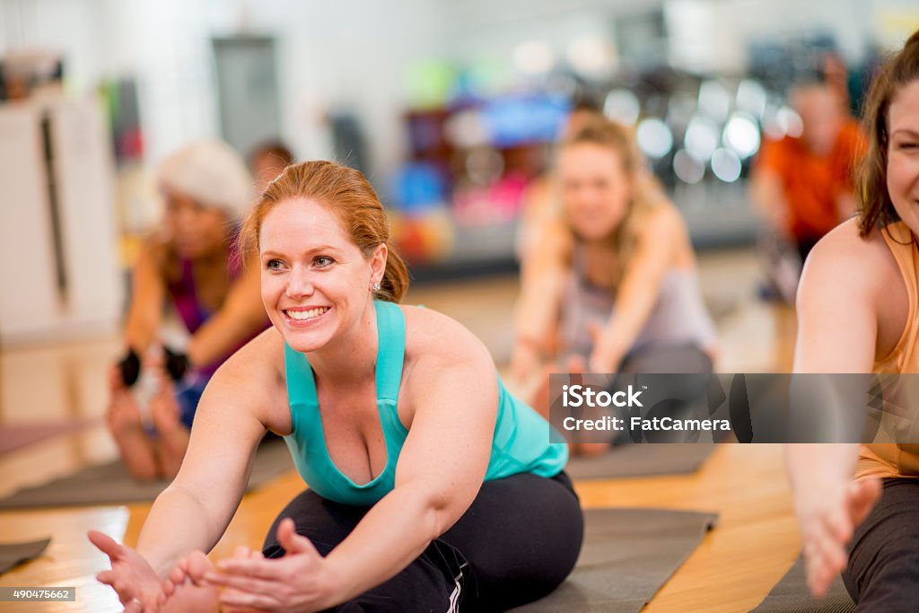 Forward Fold in Yoga Fitness Class A group of individuals are together in a gym studio stretching during a yoga class. They are sitting on their mats and have their arms outstretches in front of them in a forward fold position. Yoga Class Stock Photo