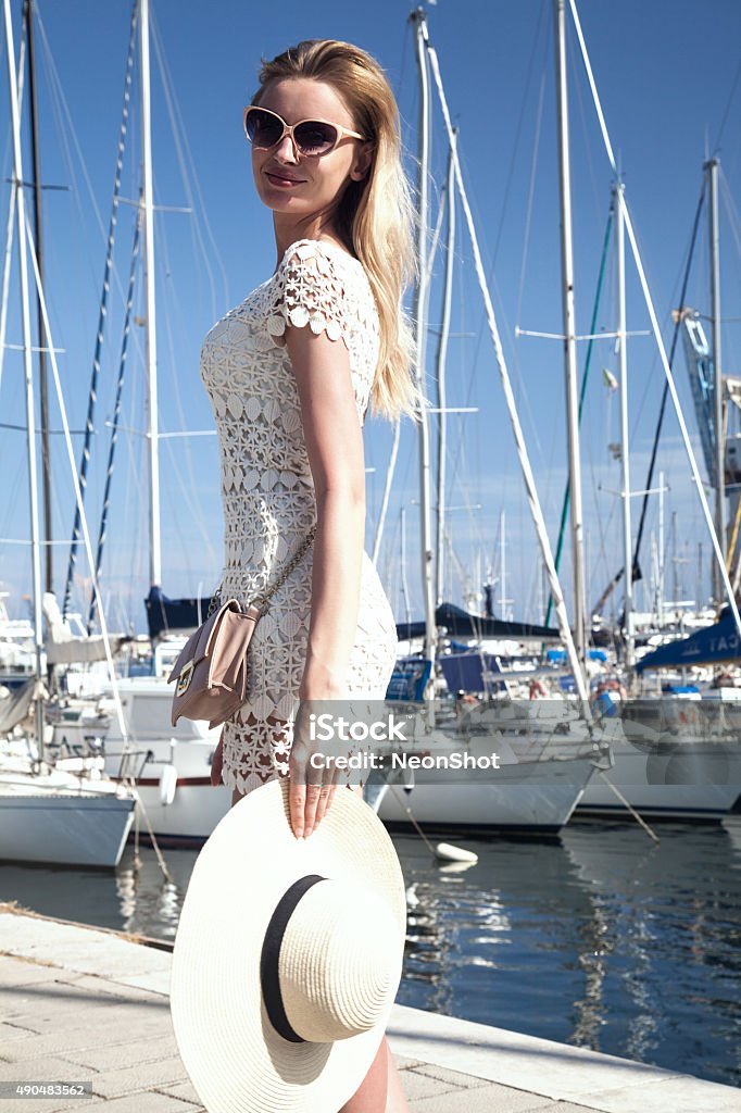 Smiling blonde lady on vacation. Elegant beautiful blonde lady posing over boats. Summer photo. Attractive smiling woman in fashionable dress and sunglasses. Blue sky. Outdoor photo. 2015 Stock Photo