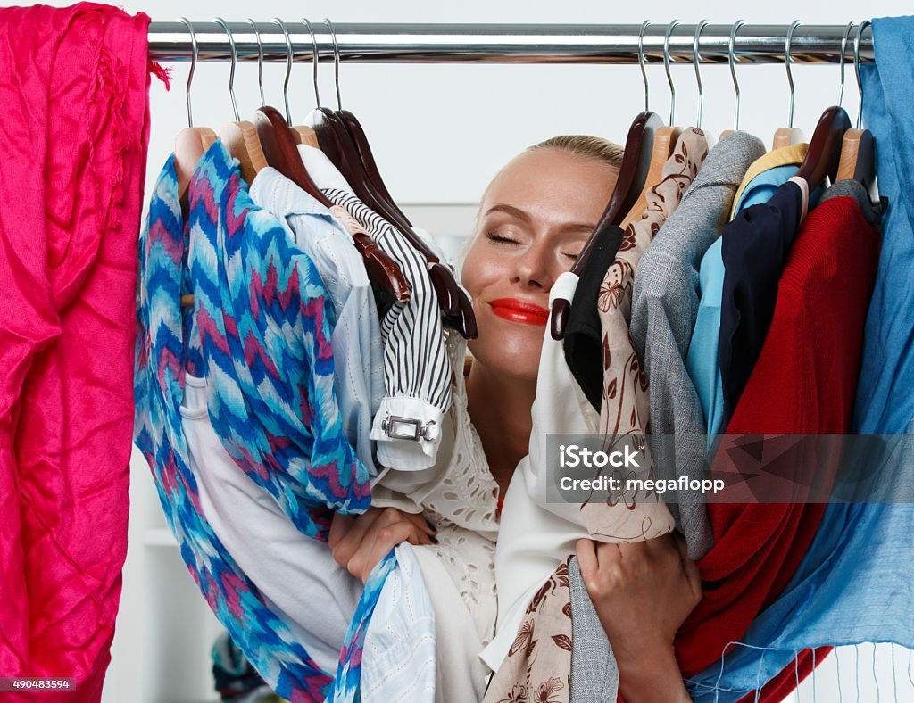 Beautiful smiling blonde woman standing inside wardrobe rack Beautiful smiling blonde woman standing inside wardrobe rack full of clothes happy to have it. Shopping and consumerism or stylist concept. Nothing to wear and hard to decide concept Change Stock Photo