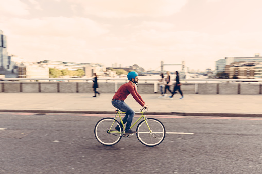 Hipster man cycling on London bridge with Thames river and Tower Bridge on background. He is riding a fixed gear bike and wearing blue jeans and a red sweater. Panning technique.
