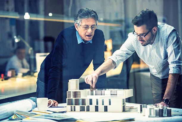 Two architects discussing new project Young architect explaining project plan to senior coworker in the office with the help of an architectural model, shot through glass. architectural model photos stock pictures, royalty-free photos & images