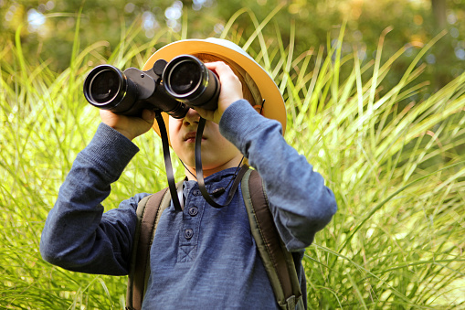This is a close-up image of a 7 year old boy dressed in blue and wearing a safari hat and a backpack, who is looking through big binoculars as he is exploring nature. The child is standing in front of a tall green grass background as he is looking sideward and holding binoculars in such a way that his face is covered.