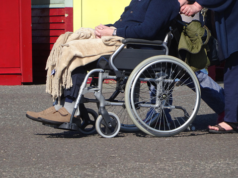 Photo showing an old disabled man being pushed along a pathway in his wheelchair, with a blanket to keep him warm.