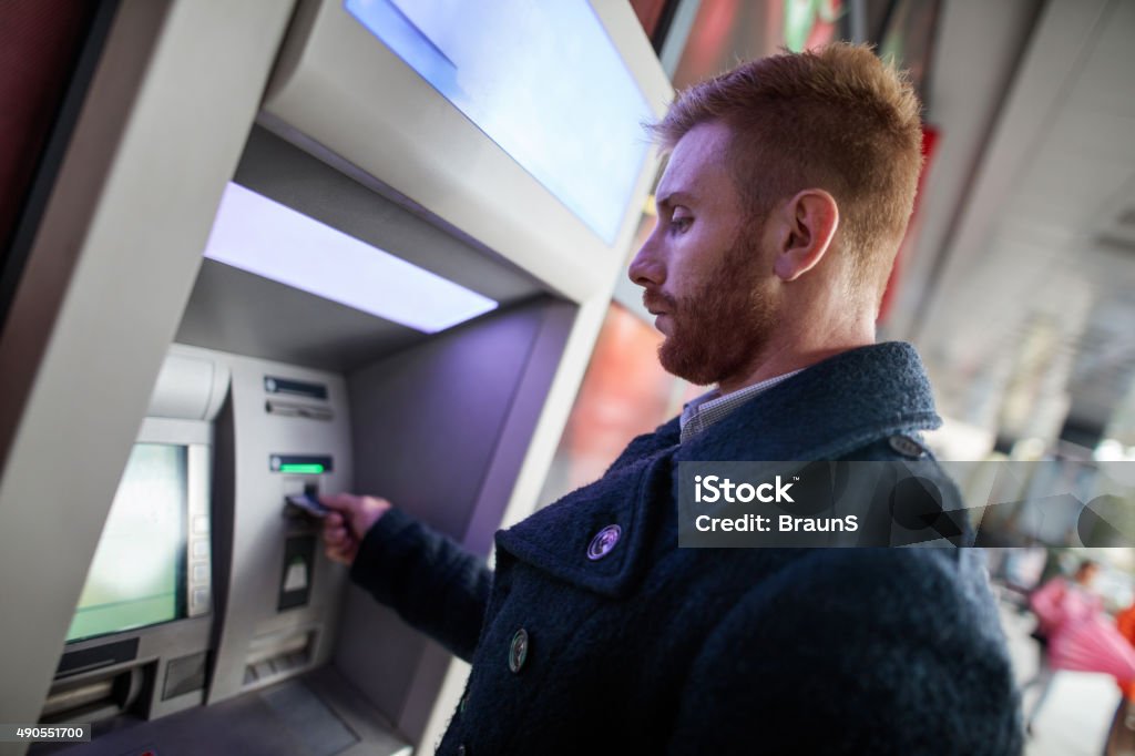 Young redhead businessman withdrawing money from ATM machine. Young businessman using cash machine in the city. ATM Stock Photo