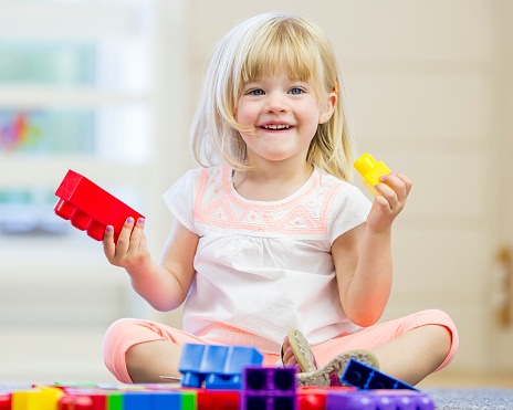 A little girl is sitting on the floor at preschool and is happily playing with colorful plastic block blocks .