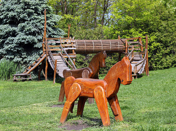 Two young girls are riding a wooden horse in an outdoor setting. The girl in front, with a focused and joyful expression, is holding onto the reins, while the girl behind her holds on tightly, smiling brightly at the camera. The wooden horse is simple and rustic, blending perfectly with the natural surroundings. The background is filled with green foliage and a hint of a wooden fence, suggesting they are in a garden or park. This image captures a moment of shared fun, adventure, and sisterly or friendly bonding in a serene outdoor environment.