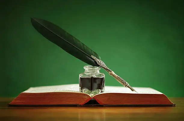 Quill pen and inkwell resting on an old book with green background concept for literature, writing, author and history
