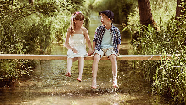 Girl in a white dress and boy with a blue hat sitting on a ledge above a small stream. They are holding hands and talking. The boy is dipping his feet in the stream. Lots of green foliage in the background.