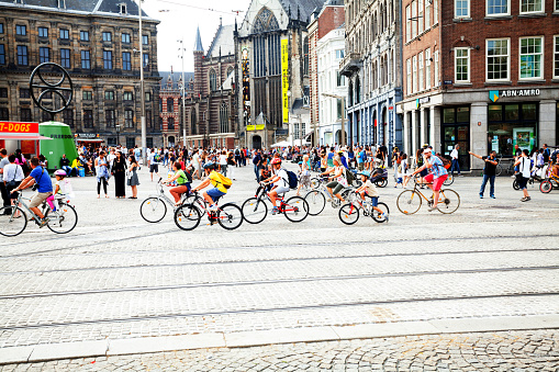 Amsterdam, The Netherlands - August 9, 2015: Many kids are cycling in street Dam in Amsterdam. A man is cycling ahead. In background are buildings and square with royal palace. Summertime shot,