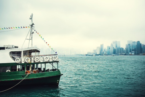 A ferry floats in the foreground of an image of Victoria Harbour, downtown Hong Kong in the background, shrouded in fog.  The ferry is a common means of transportation between Kowloon and Hong Kong.  Horizontal image with copy space.