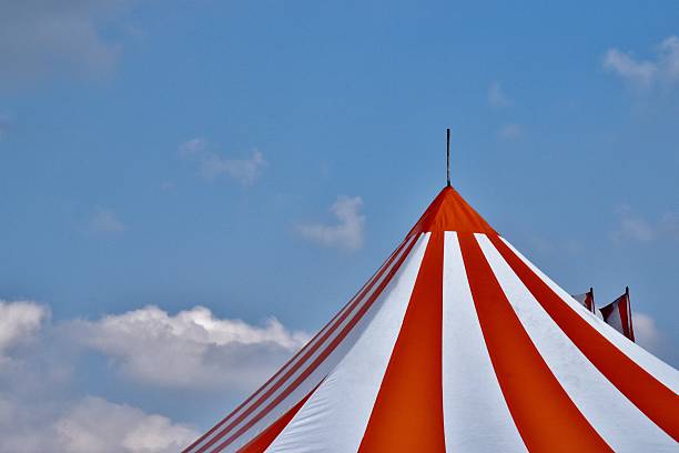 The Big Top An orange and white striped circus tent photographed under a blue summer sky, striped flags flying in the background. midway fair stock pictures, royalty-free photos & images