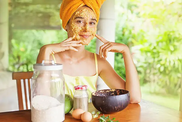 Portrait of a happy caucasian woman using natural secrets to do a skin care (ingredients are on a table: oats, flour, eggs, leaves) touching edge of her face by fingers