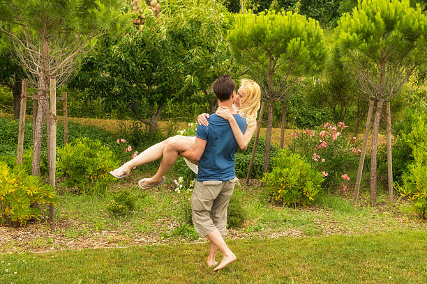 Young couple, man and woman in love,  in the garden. Man holding woman in his hands and lifting her up happily. Summer time. Women has long blond hair, white short skirt.