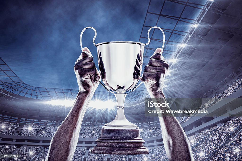 Composite image of cheering rugby player with cup Cropped hand of athlete holding trophy against football stadium Soccer Stock Photo