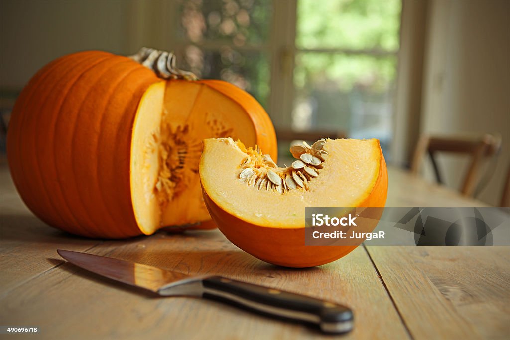Cutting a Slice of Pumpkin in a Kitchen A big orange pumpkin on a kitchen table. A slice of the squash vegetable is cut off and laying next to the pumpkin, as is the kitchen knife, in preparation of a pumpkin soup. There are no people in this image. Pumpkin Stock Photo