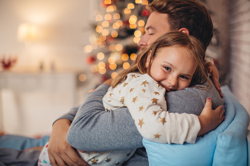 Father and his cute daughter in front of christmas tree, lying on bed. Girl embracing daddy.