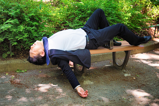 Passed out man in Tokyo park daytime profile full length. He is lying down on a  park bench, dressed in a suit.