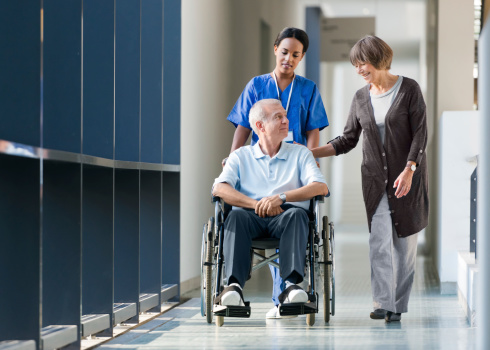 Senior couple with man in wheelchair and a nurse walking a hospital corridor and talking. 
