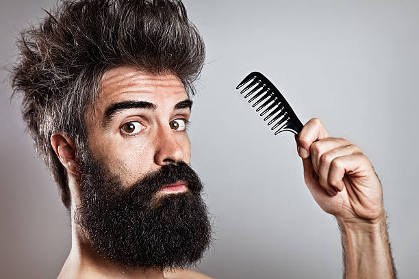 Messy haired bearded man holding a comb looking at came Close-up of a long bearded man with crazy hairstyle and doubtful expression staring at camera holding a comb in his hand bushy stock pictures, royalty-free photos & images