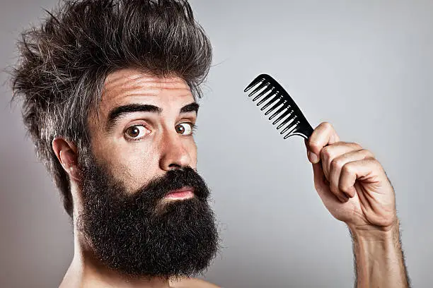 Close-up of a long bearded man with crazy hairstyle and doubtful expression staring at camera holding a comb in his hand