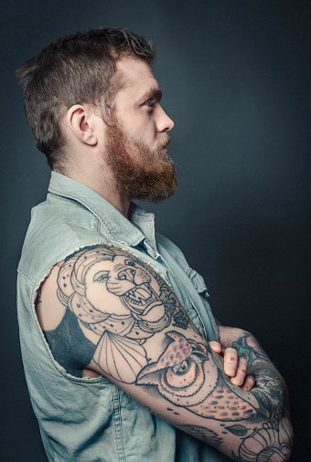 A photo of fashionable tattooed man. Side view of confident male against grey background. Hipster is with animal tattoos on arm.