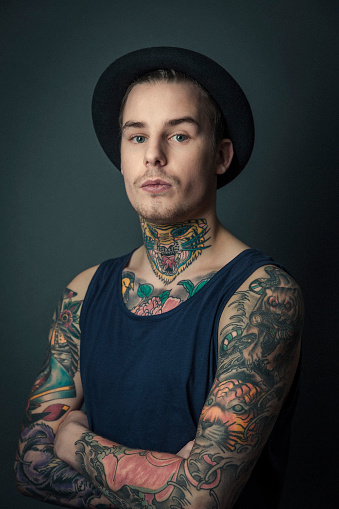 A photo of young tattooed man standing arms crossed. Portrait of stylish male against grey background. Confident hipster is with colourful tattoos.