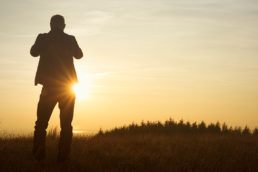 silhouette of a Man stood in countryside taking a picture with a mobile phone of an Autumn sunset.
