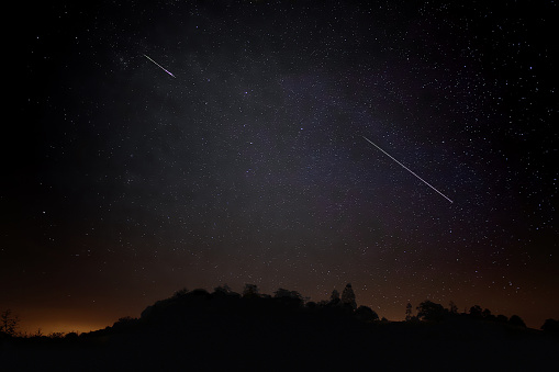 Two Meteors cross the sky, ground light and silhouette of hill landscape. Persid Annual Meteor Shower August.