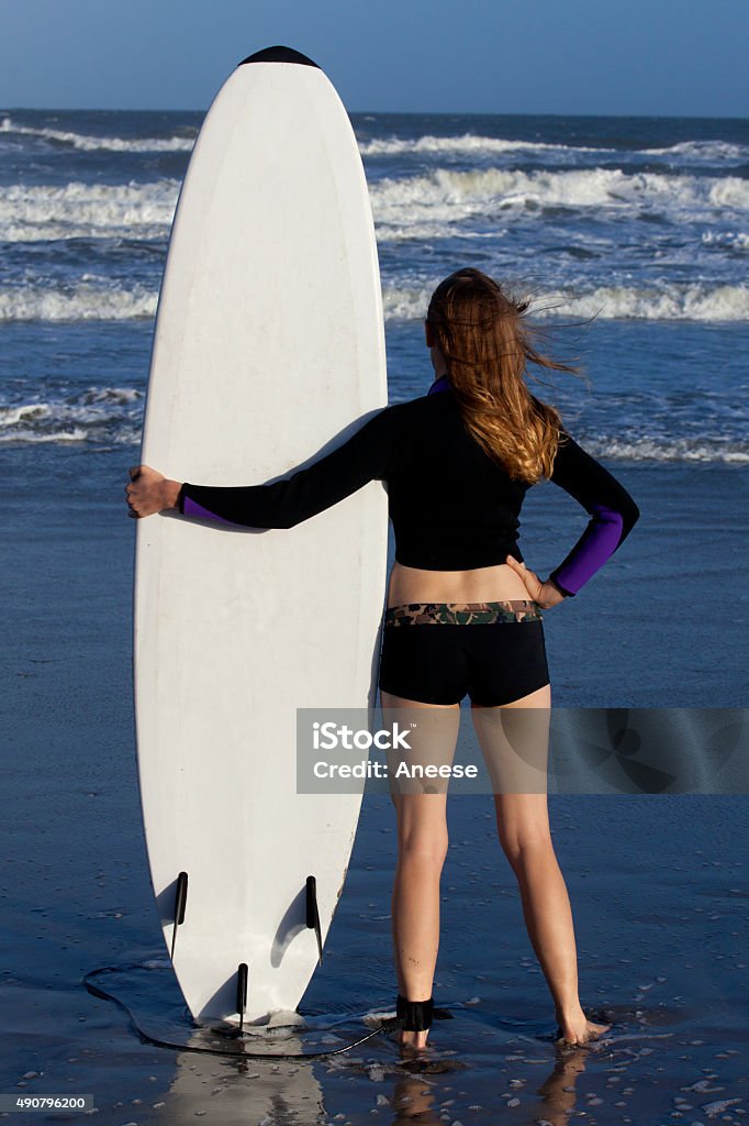 Woman with Surfboard Woman in bikini and wetsuit top, standing on beach looking at the surf on the ocean while holding a white surfboard. 20-29 Years Stock Photo