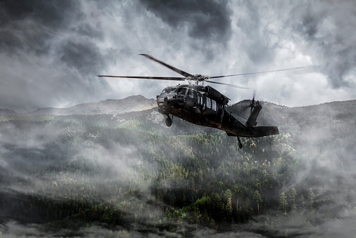 An army helicopter flies over foggy mountains.