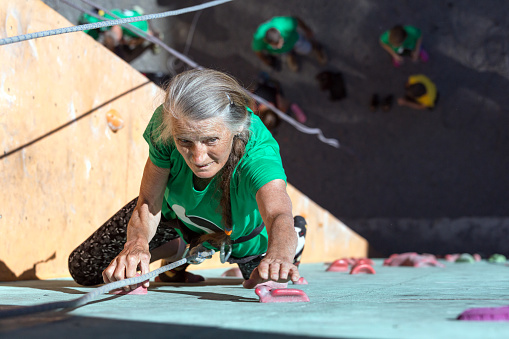 Elderly Female Demonstrates Excellent Physical and Moral Abilities Ascending Vertical Climbing Wall Group of Climbers Staying Below on Ground