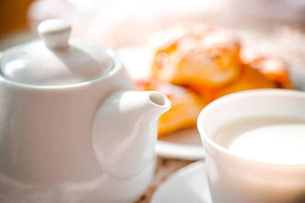 Cup of milk and tea pot with served freshly baked sweet fruit danish desserts pastries in blurred background.