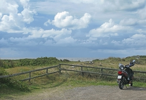 Motorcycle parked on gravel road by wooden fence in natural heath landscape at Skrea Strand on a sunny day with dark clouds in Falkenberg, Sweden.
