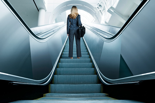 A rear view of a businesswoman, carrying a shoulder bag, riding up an escalator in an airport or office interior.